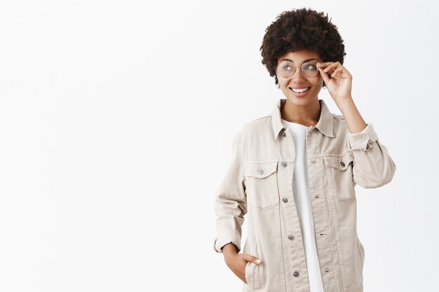 Retrato de linda adorable mujer adulta afroamericana con camisa beige y gafas tocando el borde de las gafas mientras sonríe con alegría y mira a la izquierda mirando las tiendas que pasan caminando por la ciudad