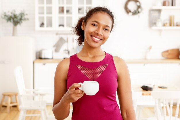 Retrato de linda chica afroamericana con cabello recogido posando en el interior de la cocina con una taza de té. Atractiva mujer de piel oscura feliz tomando café con una gran sonrisa