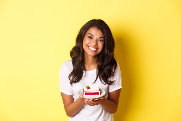 Retrato de linda chica afroamericana, sosteniendo un trozo de pastel y sonriendo, de pie sobre fondo amarillo