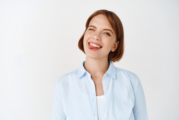 Retrato de una linda chica alegre con el pelo corto guiñando un ojo y mostrando la lengua emocionada divirtiéndose de pie en blusa sobre fondo blanco.