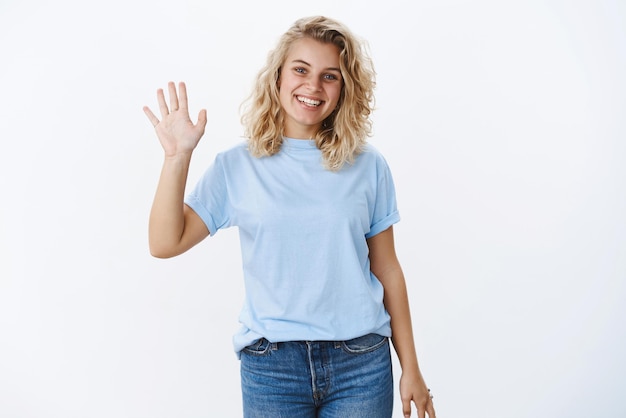 Retrato de linda chica amable y positiva con cabello rubio y ojos azules, sonriendo agradable como agitando la palma en gesto de saludo o hola, siendo acogedor y agradable sobre una pared blanca