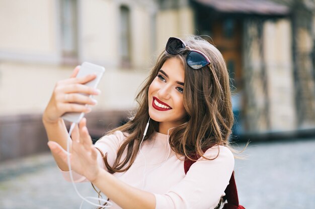 Retrato de linda chica con cabello largo y labios vinosos haciendo selfie en la calle en la ciudad. Viste camisa blanca, sonriendo.