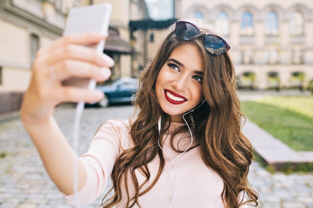 Retrato de linda chica con cabello largo y sonrisa blanca como la nieve haciendo selfie en la calle en la ciudad. Lleva labios vinosos, sonriendo.