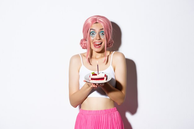 Retrato de linda chica lamiendo los labios sosteniendo un delicioso pastel, celebrando un cumpleaños, vistiendo una peluca rosa y un traje brillante para la fiesta.