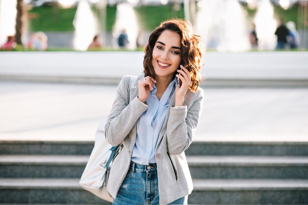 Retrato de linda chica morena con pelo corto caminando en la ciudad. Viste camisa, jeans, chaqueta y bolso. Ella está hablando por teléfono y sonriendo a la cámara.