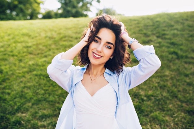 Retrato de linda chica morena con pelo corto posando para la cámara en el parque. Viste camiseta blanca y camisa azul.