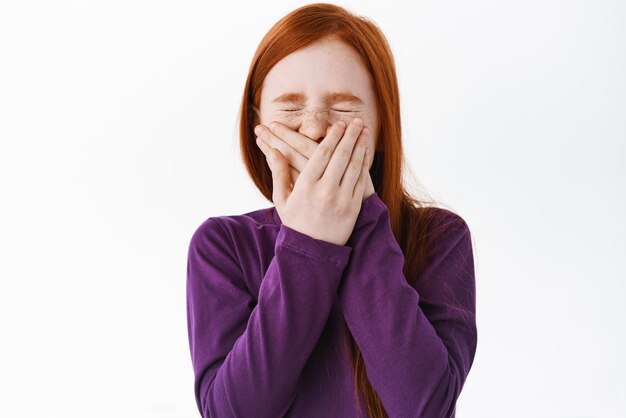 Retrato de una linda chica pelirroja estornudando cubriendo su boca y nariz arrugada de pie sobre fondo blanco Niño escondiendo su sonrisa riendo y bromeando