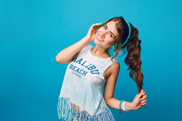 Retrato de linda chica con pelo largo y rizado en cola sobre fondo azul. Viste camiseta blanca, pantalones cortos y escucha música con auriculares azules.