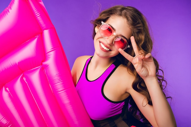 Retrato de linda chica con pelo largo y rizado en gafas de sol rosas sonriendo a la cámara sobre fondo púrpura en estudio. Viste traje de baño, sostiene un colchón de aire rosa y muestra un cartel genial.