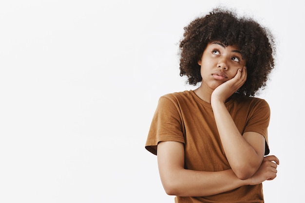 Retrato de una linda chica de piel oscura espaciada con peinado afro en camiseta marrón con la cara inclinada en la palma y mirando hacia arriba mientras usa la imaginación