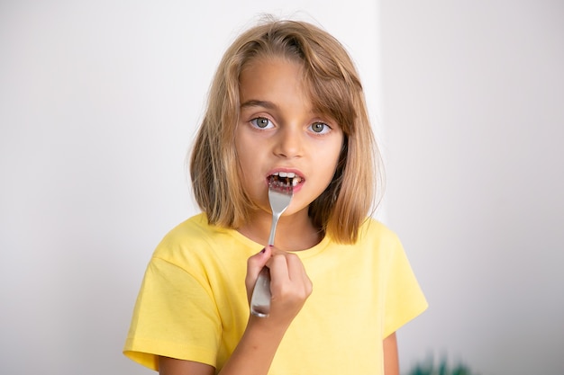 Retrato de linda chica rubia comiendo pastel con tenedor. Niño bastante caucásico de pie, comiendo. Concepto de infancia, celebración y vacaciones.