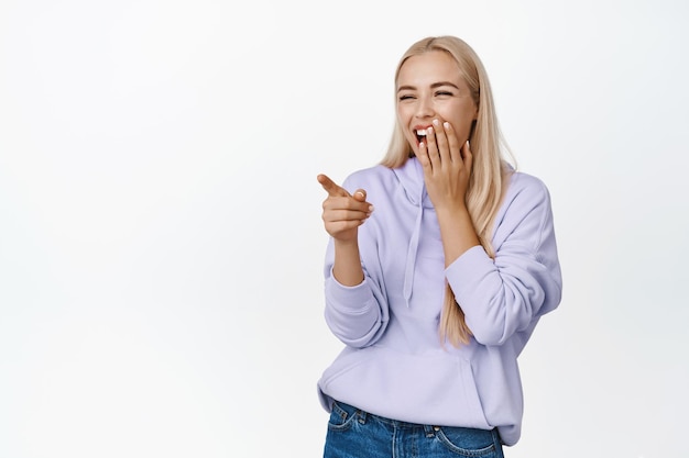 Retrato de una linda chica rubia que se ríe de algo divertido que señala con el dedo algo gracioso y se ríe de pie sobre un fondo blanco