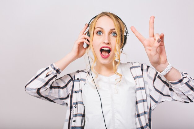 Retrato de linda chica rubia rizada en auriculares escuchando música con cara de sorpresa y bailando. Encantadora dama de ojos azules con la boca abierta muestra el signo de la paz divirtiéndose y disfruta de su canción favorita.