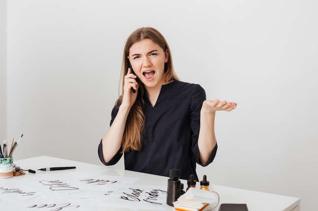 Retrato de una linda chica sentada en el escritorio blanco y hablando por su teléfono celular mientras mira sorprendida en la cámara aislada