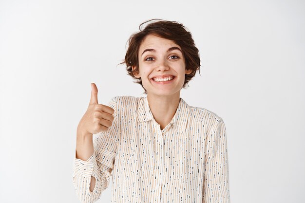 Retrato de linda chica solidaria mostrando los pulgares hacia arriba y sonriendo orgullosa, felicitándote, mostrando bien hecho, excelente gesto, de pie satisfecho en la pared blanca