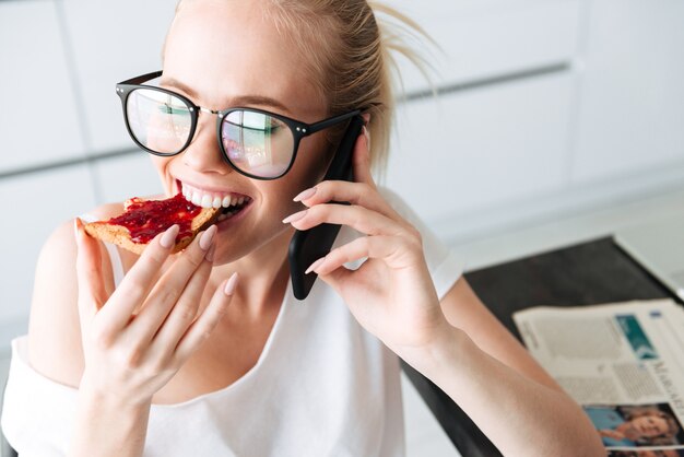 Retrato de linda dama disfrutando de pan con mermelada y hablando por teléfono inteligente en la cocina