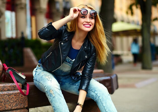 Retrato de linda divertida moderna sexy urbano joven elegante mujer sonriente modelo de niña en tela moderna brillante al aire libre sentado en el parque en jeans en un banco en gafas