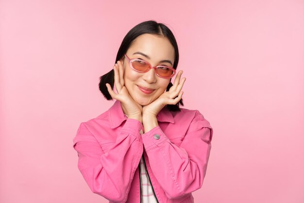 Retrato de una linda y elegante chica asiática sonriendo y tocando la cara mirando hacia arriba con una mirada pensativa de ensueño de pie sobre un fondo rosa