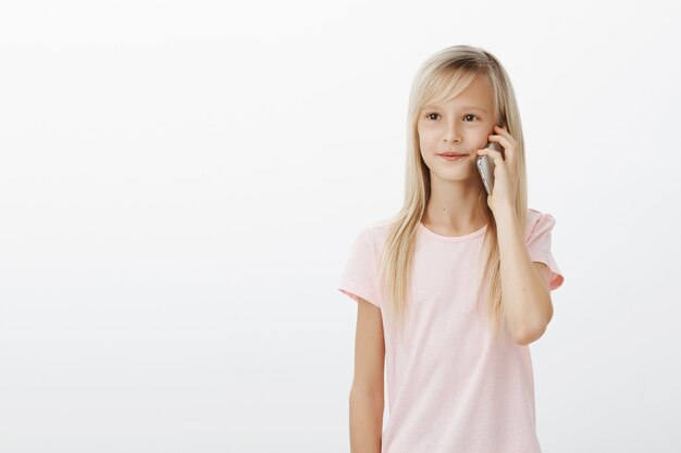 Retrato de linda hija joven con cabello rubio, sosteniendo el teléfono inteligente cerca de la oreja y mirando a un lado con expresión de satisfacción enfocada, hablando con su hermano