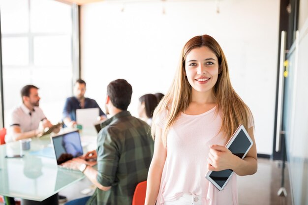 Foto gratuita retrato de una linda joven mujer de negocios con tableta digital que parece segura en la sala de reuniones con colegas en la parte de atrás