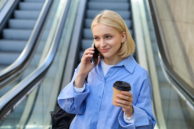 Foto gratuita retrato de una linda joven rubia estudiante con una taza de café responde a una llamada telefónica de pie cerca