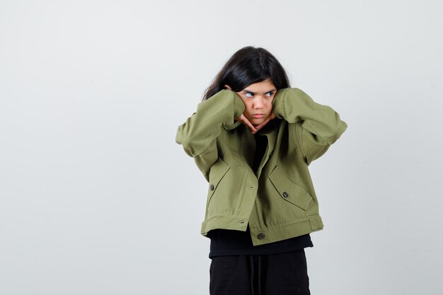 Retrato de linda jovencita enfurruñada con las mejillas apoyadas en las manos, mirando a otro lado con chaqueta verde militar y mirando pensativa vista frontal