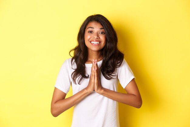 Retrato de linda mujer afroamericana, sonriendo y tomados de la mano en oración, diciendo gracias, sintiéndose agradecido, de pie sobre fondo amarillo.