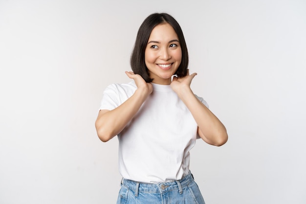 Retrato de linda mujer asiática hermosa tocando su nuevo corte de pelo corto que muestra el peinado sonriendo feliz a la cámara de pie sobre fondo blanco.