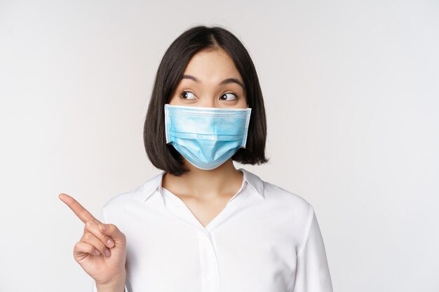 Retrato de una linda mujer asiática con mascarilla médica protección contra el coronavirus apuntando con el dedo a la izquierda y mirando intrigada por el espacio vacío de la copia de fondo blanco