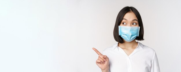 Retrato de una linda mujer asiática con mascarilla médica protección contra el coronavirus apuntando con el dedo a la izquierda y mirando intrigada por el espacio vacío de la copia de fondo blanco