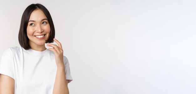 Retrato de una linda mujer coqueta riendo y sonriendo mirando a un lado pensando reflexivamente o imaginando algo de pie en una camiseta blanca sobre el fondo del estudio