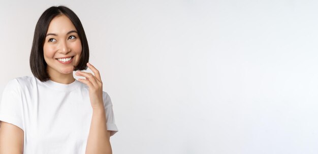 Retrato de una linda mujer coqueta riendo y sonriendo mirando a un lado pensando reflexivamente o imaginando algo de pie en una camiseta blanca sobre el fondo del estudio