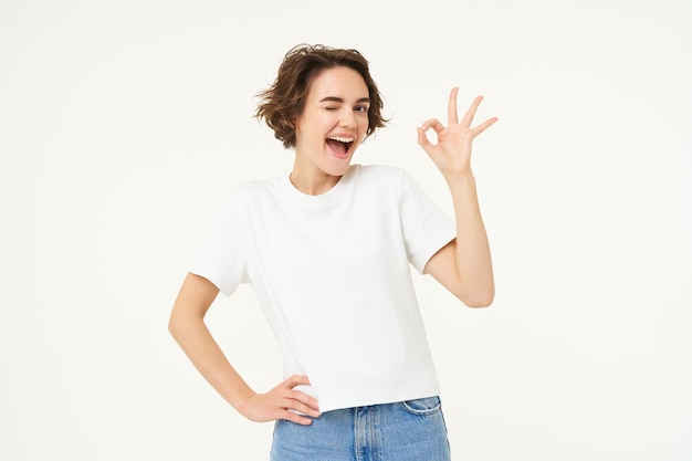 Foto gratuita el retrato de una linda mujer emocionada te guiña un ojo y muestra un gesto bien bueno posando sobre el fondo blanco del estudio