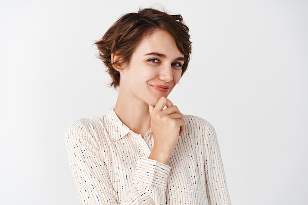 Retrato de linda mujer moderna en blusa con peinado corto, sonriendo y tocando la barbilla con expresión pensativa, teniendo una idea, mirando algo, pared blanca