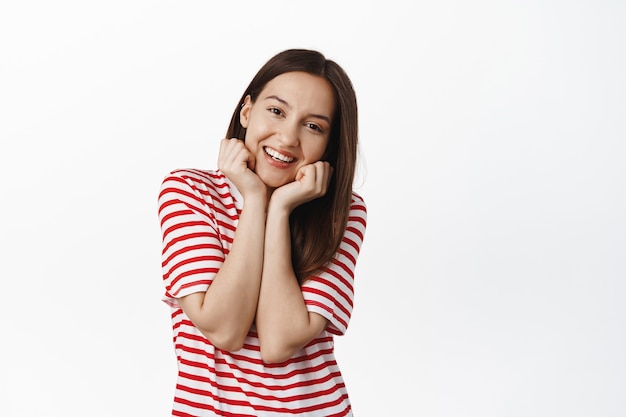 Retrato de linda mujer morena sonriendo, inclinando la cabeza y mirando con admiración, luciendo adorable, tomados de la mano cerca de la cara y sonriendo feliz, de pie contra la pared blanca