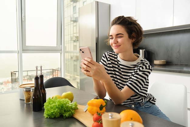 Foto gratuita retrato de una linda mujer sonriente cocinando ensalada mirando el teléfono inteligente viendo la receta de preparación de alimentos