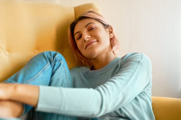 Retrato de linda niña de 20 años con anillo en la nariz y cabello rosado holgazaneando en un sillón vistiendo ropa casual con expresión facial relajada y despreocupada.