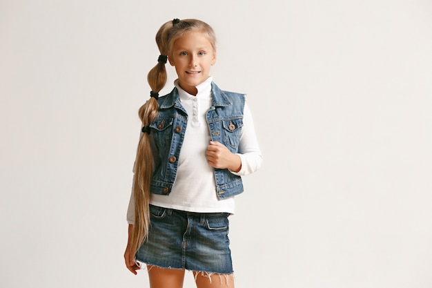 Retrato de linda niña adolescente en ropa de jeans con estilo mirando a cámara y sonriendo contra la pared blanca del estudio. Concepto de moda infantil