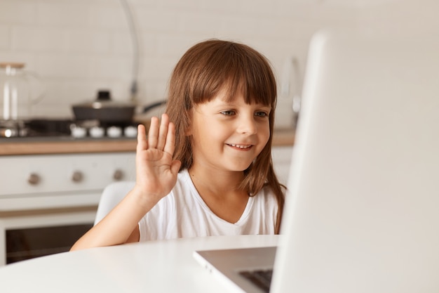 Retrato de linda niña de pelo oscuro sentada en la mesa con videollamada, mirando la pantalla del portátil y agitando la mano a la cámara web del portátil, tiene expresión positiva.