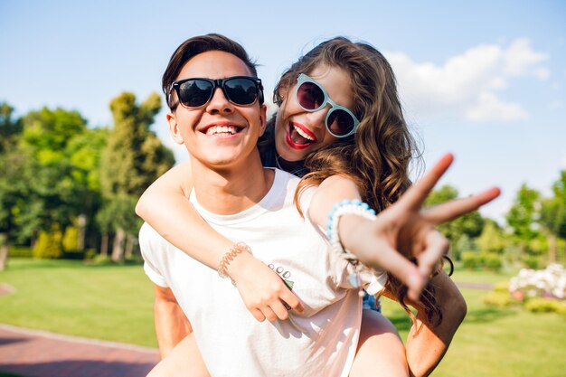 Retrato de linda pareja divirtiéndose en el parque. Chica guapa con pelo largo y rizado está montando en la espalda de un chico guapo. Llevan gafas de sol y sonríen a la cámara.