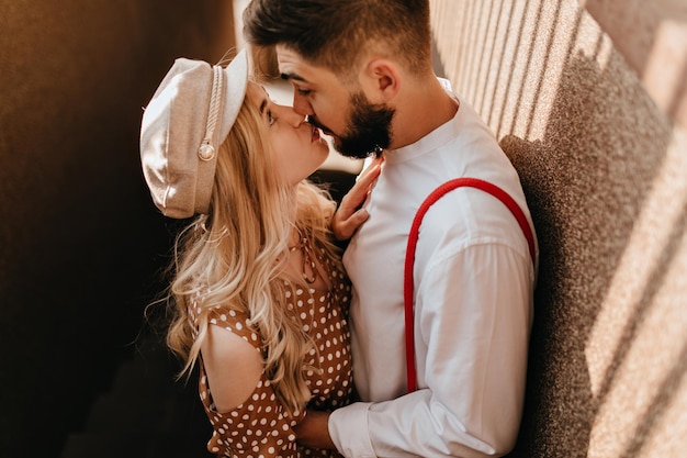 Retrato de linda pareja romántica besándose al aire libre en un día soleado Chica con gorra beige y vestido marrón mira a los ojos de su amado novio