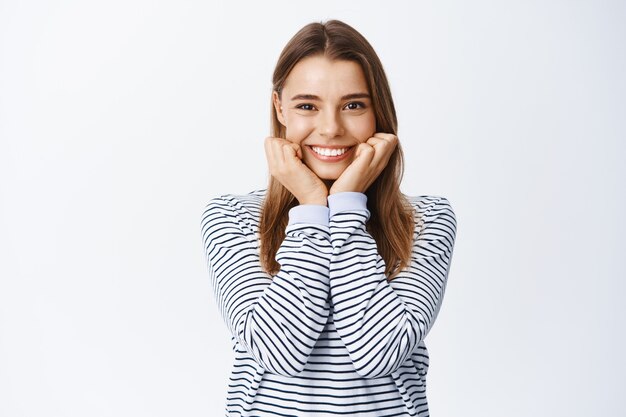 Retrato de linda y romántica chica rubia apoyada en las manos y sonriendo al frente, mirando algo hermoso, de pie asombrado contra la pared blanca