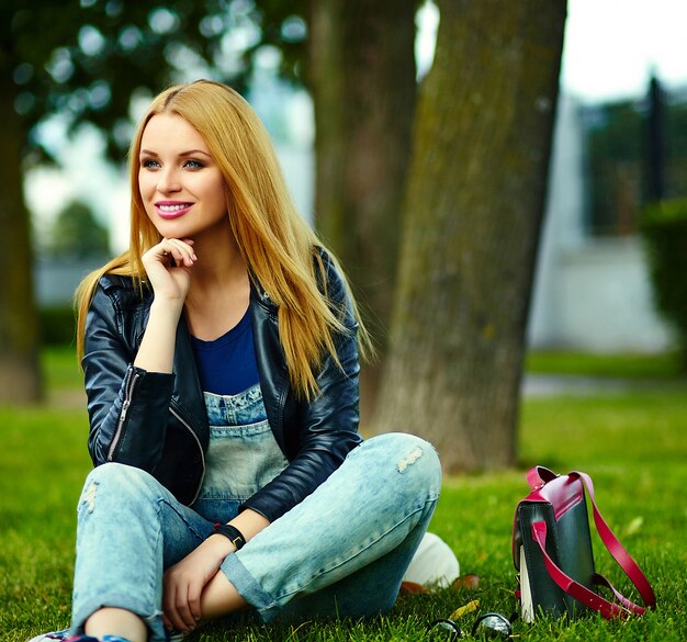 Retrato de linda rubia divertida moderna sexy urbano joven elegante mujer sonriente modelo de niña en tela moderna brillante al aire libre sentado en el parque en jeans con bolsa rosa