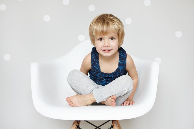 Retrato de lindo bebé caucásico con cabello rubio y grandes ojos hermosos, vestido con traje de dormir, sentado con las piernas cruzadas en una silla blanca, mirando y sonriendo, negándose a ir a la cama