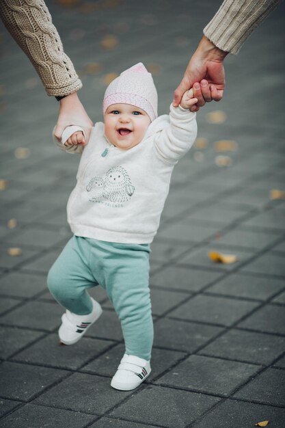 Retrato de un lindo y dulce bebé mientras camina afuera Padres sosteniendo a su hijo satisfecho con las manos mientras camina Feliz bebé sonriente con suéter blanco, pantalones verdes y zapatillas blancas haciendo pasos