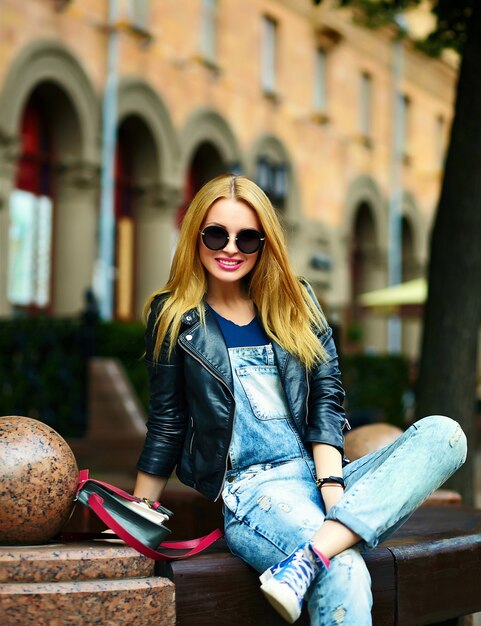 Retrato de lindo gracioso rubio moderno sexy urbano joven elegante mujer sonriente modelo de niña en tela moderna brillante al aire libre sentado en el parque en jeans en un banco en gafas