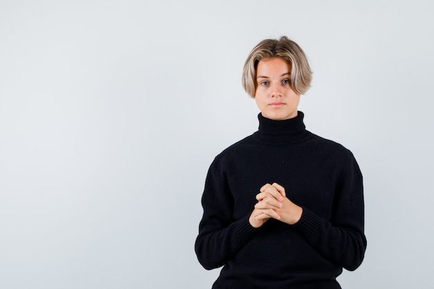 Retrato de lindo muchacho adolescente con las manos entrelazadas en suéter de cuello alto negro y mirando pensativo vista frontal