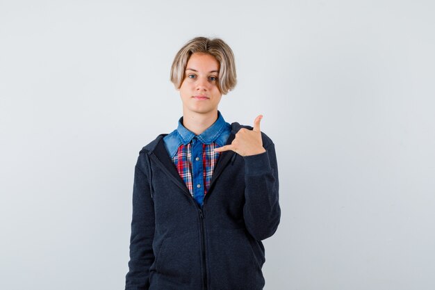 Retrato de lindo muchacho adolescente mostrando gesto de teléfono en camisa, sudadera con capucha y mirando confiado vista frontal