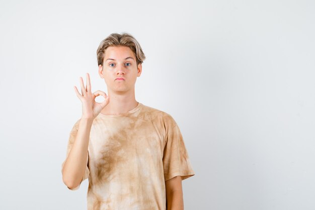 Retrato de un lindo muchacho adolescente que muestra un gesto de ok en camiseta y parece confundido vista frontal