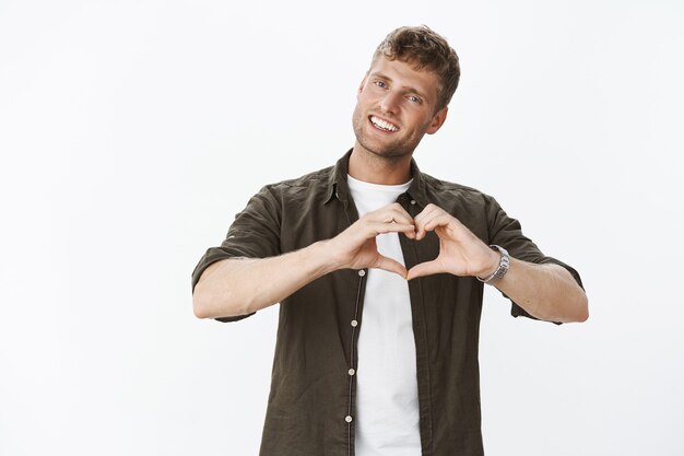 Retrato de lindo y tierno novio masculino guapo sonriendo e inclinando la cabeza mostrando el signo del corazón sobre el cuerpo, haciendo confesión de simpatía y sentimientos románticos sobre una pared gris